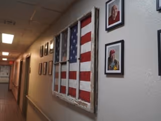 A hallway with framed pictures on the wall, including a large framed American flag and several portraits of elderly individuals.