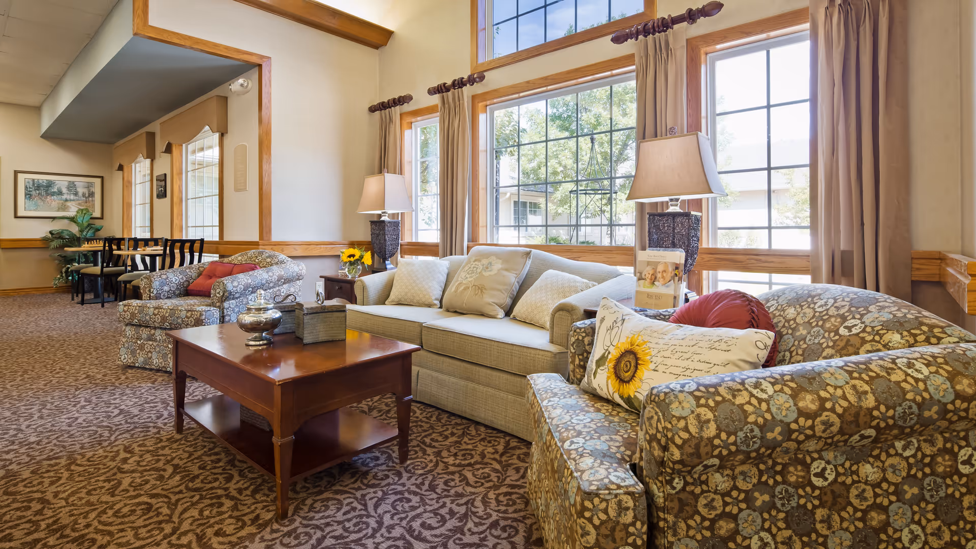 A cozy living room area in Park West Plaza with large windows letting in natural light. The room features a beige sofa with decorative pillows, two patterned armchairs with cushions, a wooden coffee table with decorative items, two table lamps, and a small table with chairs in the background near framed artwork and plants.