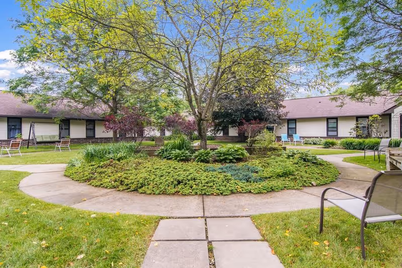 A circular garden with various green plants and a tree in the center, surrounded by a concrete walkway. Single-story buildings with beige siding and brown roofs encircle the garden. Several chairs and benches are placed along the walkway, and the sky is clear with a few clouds.