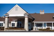 Exterior view of a single-story assisted living facility building with a covered entrance supported by stone pillars, beige siding, and a brown shingled roof under a clear blue sky.