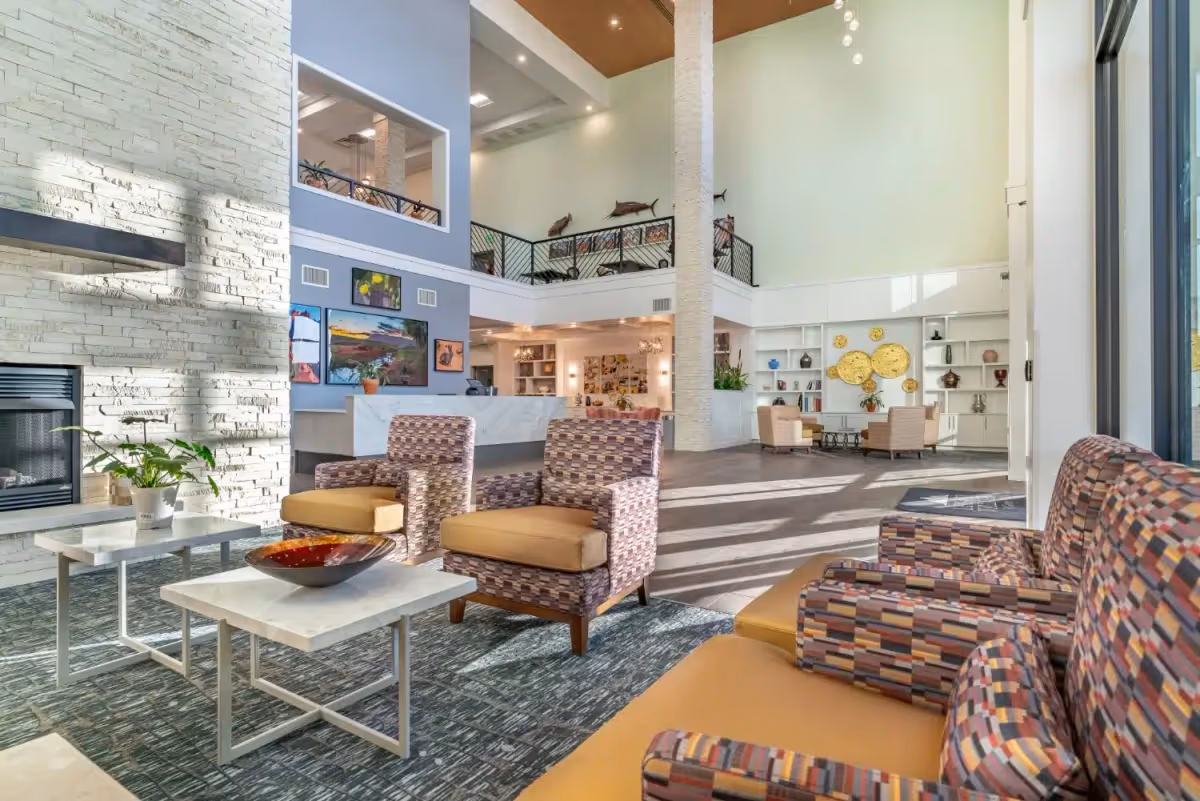 A spacious and well-lit living room area in a senior living facility featuring patterned armchairs with mustard cushions, white stone fireplace, modern white tables with decorative items, and a high ceiling with a balcony railing above. The room has large windows allowing natural light to fill the space, with artwork and decorative elements on the walls and shelves.