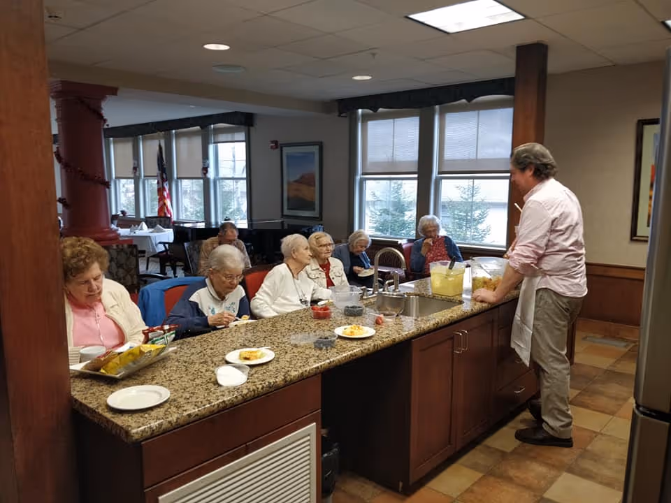 A group of elderly people sitting at a granite countertop in a senior living facility, eating and interacting with a man standing behind the counter who is serving drinks. The room has large windows with blinds, wooden cabinetry, and a tiled floor.