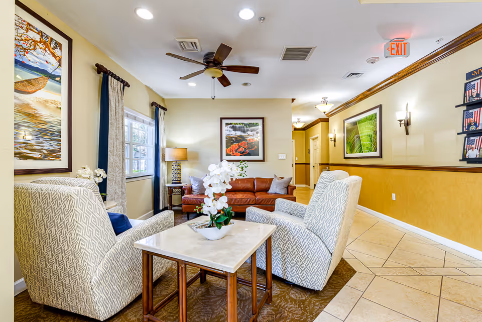 Bright senior living lounge with patterned armchairs and a leather sofa arranged around a coffee table, framed artwork on the walls and a ceiling fan overhead.
