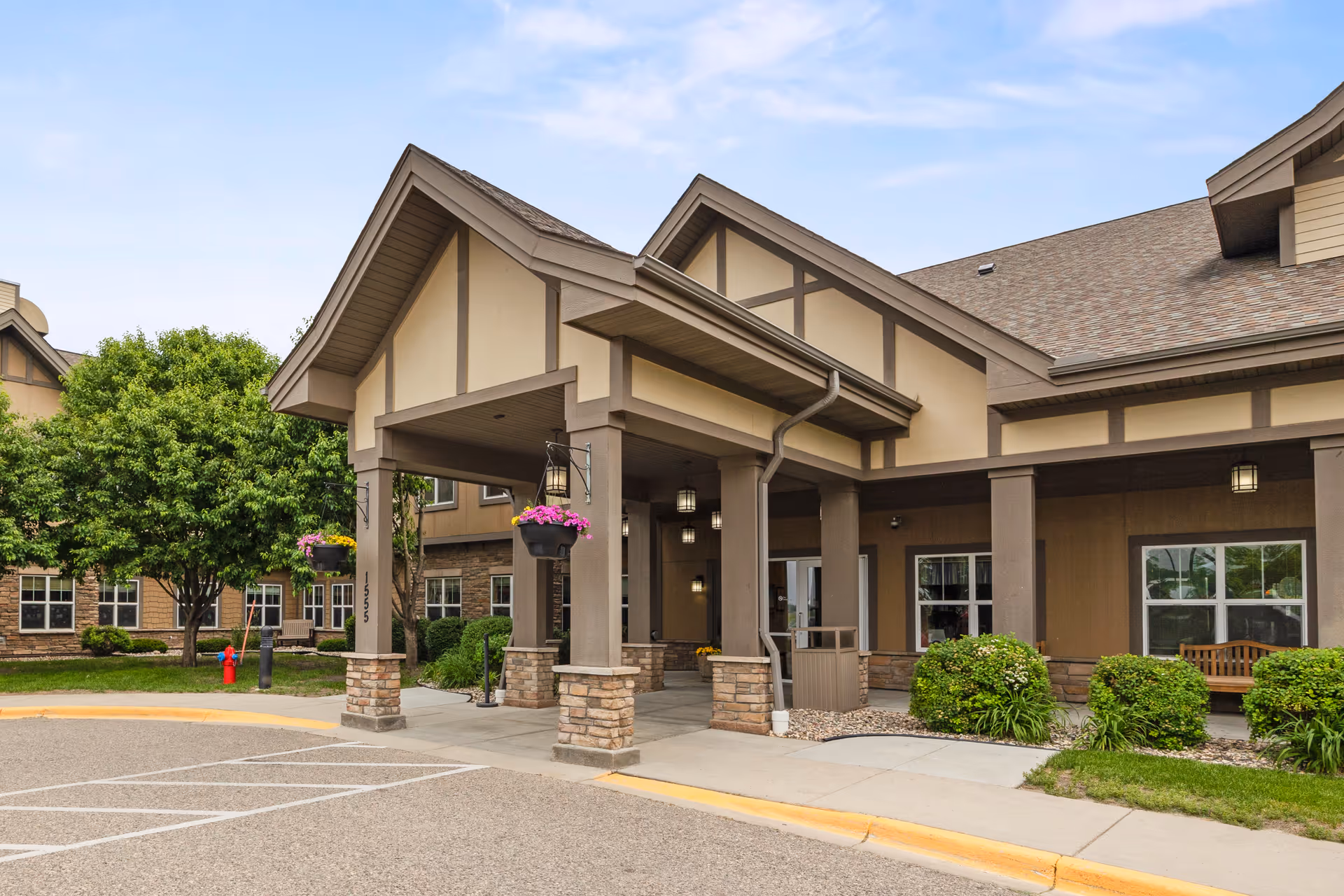 Exterior view of the entrance to a senior living facility with a covered drop-off area supported by columns with stone bases. There are hanging flower pots with pink flowers, green bushes, a tree, and a bench near the building. The sky is clear with some clouds.