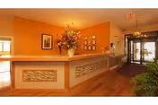 Reception area of a senior living facility with a wooden front desk decorated with a large floral arrangement. The walls are painted orange and adorned with framed pictures. There is a chandelier hanging from the ceiling and a glass entrance door in the background.