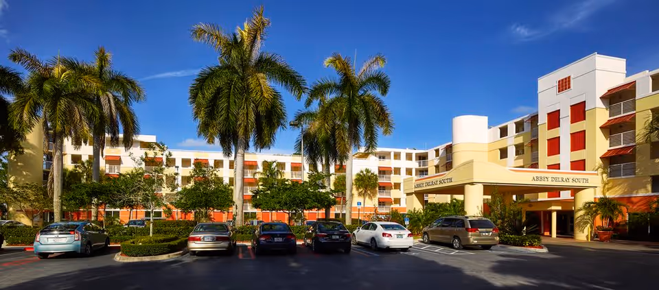 Exterior view of Abbey Delray South senior living facility with a parking lot in front, several cars parked, tall palm trees, and a clear blue sky.