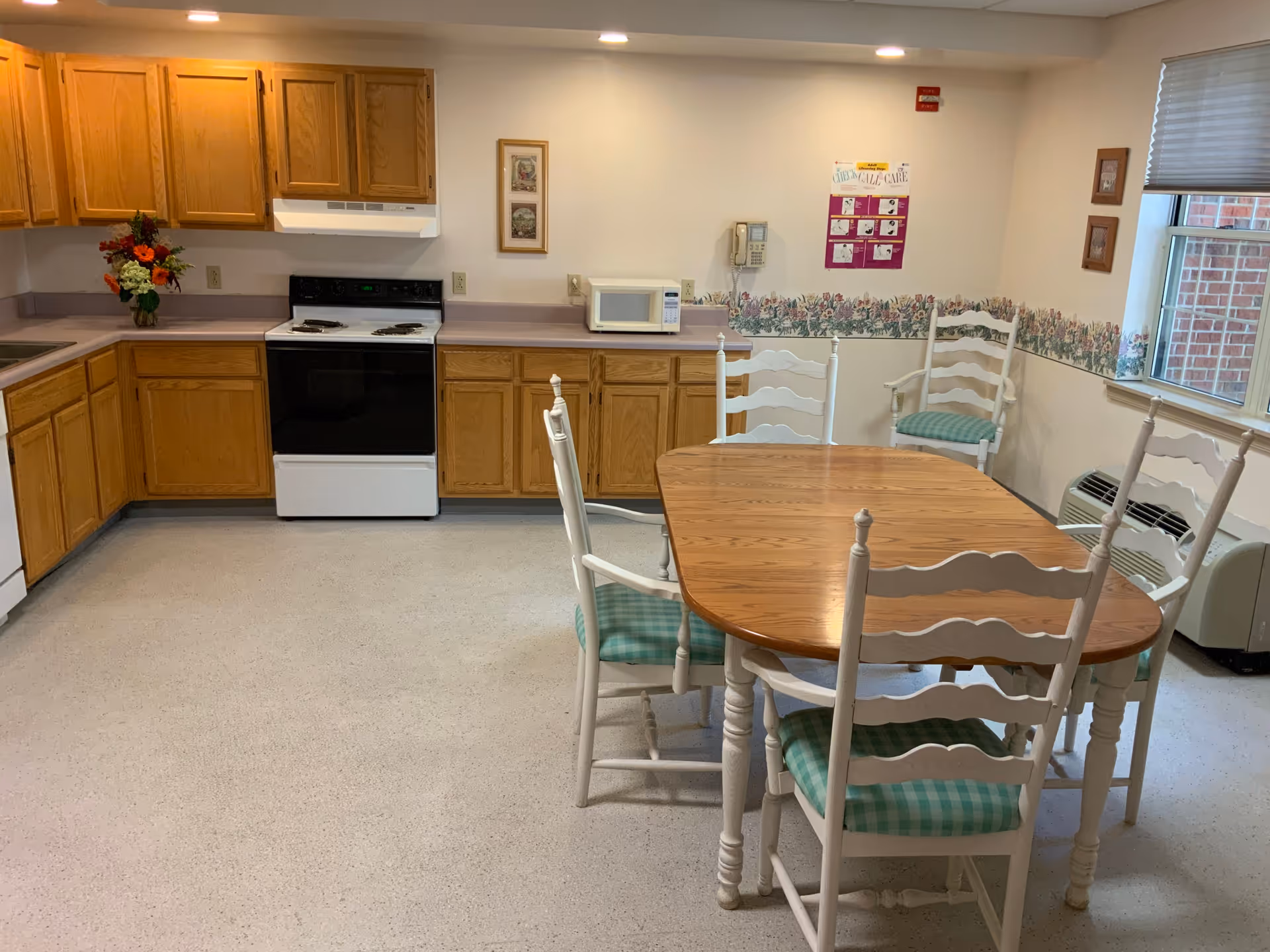 A kitchen and dining area with wooden cabinets, a white stove, microwave, and a wooden dining table with six white chairs that have green checkered cushions. There is a window with blinds, floral wallpaper border, and a wall-mounted phone.