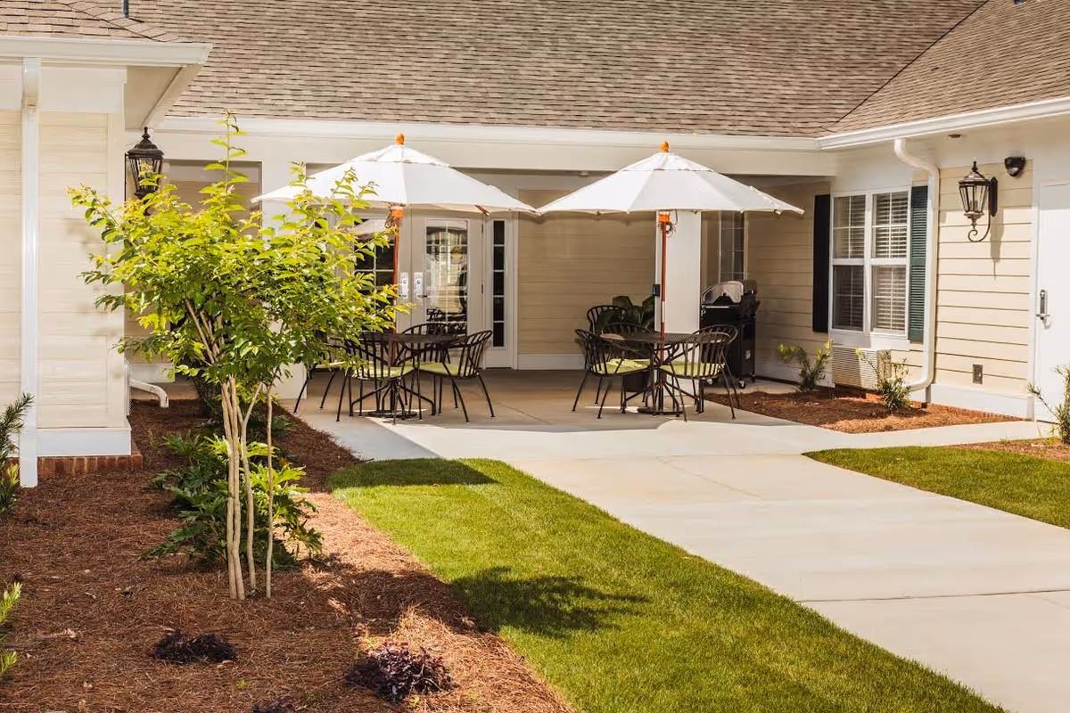 Outdoor patio area at a senior living facility with two tables, each shaded by a white umbrella, surrounded by chairs. The patio is adjacent to a beige building with windows and a door, and there is a small tree and landscaped garden beds with mulch and plants nearby.