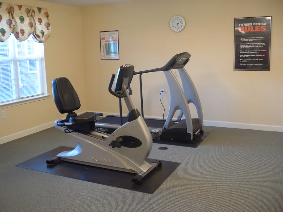 A small fitness center room with a recumbent exercise bike and a treadmill on black mats. The room has beige walls, a window with a curtain featuring a hot air balloon pattern, a wall clock, and two framed posters including one titled 'Fitness Center Rules'.