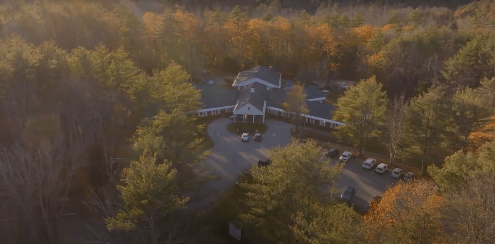 Aerial view of a single-story building surrounded by dense trees with autumn foliage. The building has a circular driveway with several parked cars. The scene is bathed in warm sunlight.