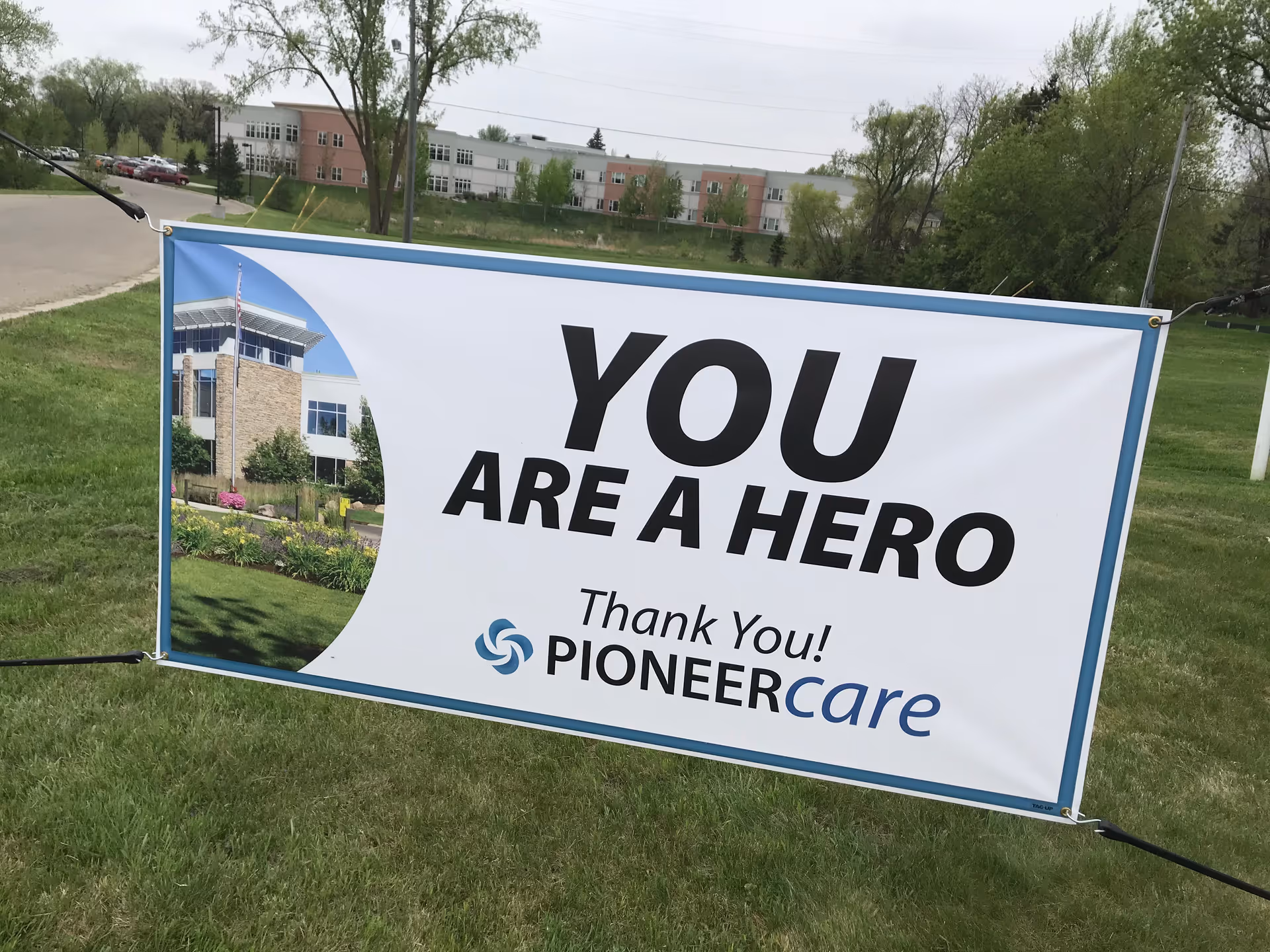 A large outdoor banner on a grassy area with trees and a building in the background. The banner reads 'YOU ARE A HERO Thank You! PIONEERcare' with a small image of a building on the left side.