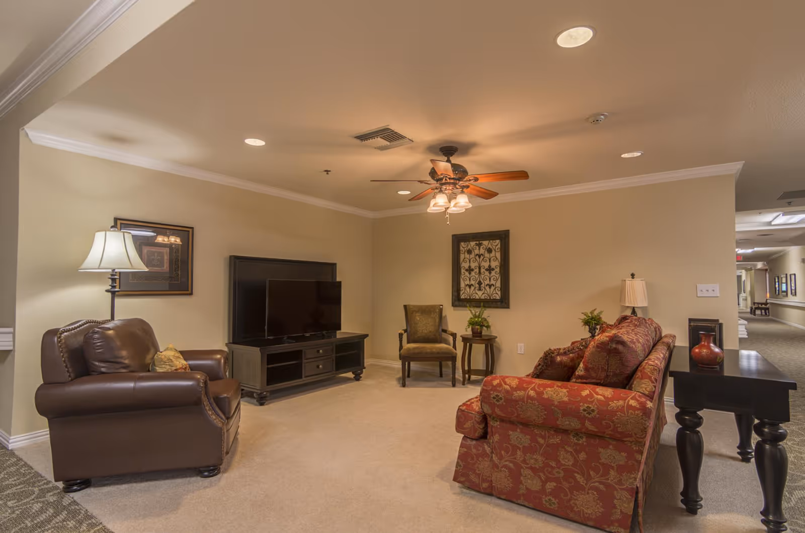 A cozy living room area in an assisted living facility with a brown leather armchair, a red patterned sofa, a green upholstered chair, a TV on a black stand, a ceiling fan with lights, framed wall art, and side tables with lamps and plants.