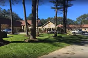 Exterior view of Alpine Nursing Home building with a driveway, several parked cars, tall trees, and a well-maintained lawn under a clear blue sky.