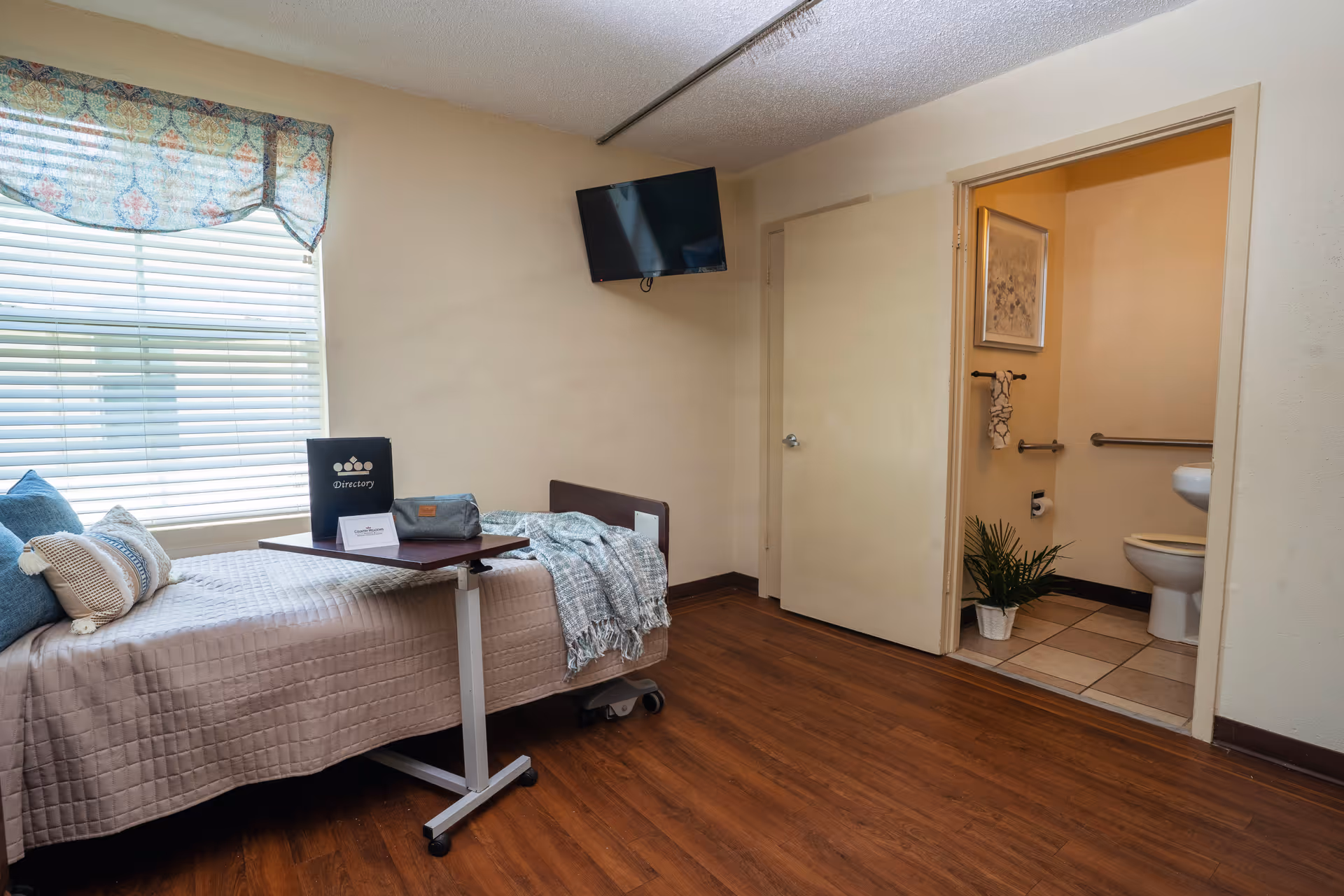Private nursing facility room with a single bed, overbed table, wall-mounted TV and an open doorway to an accessible bathroom.