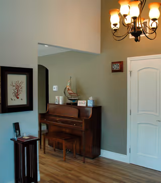 Interior view of a room with a wooden upright piano and matching bench against a beige wall. Above the piano is a decorative model sailboat and some small framed pictures. To the right is a white door with a small framed artwork above it. A chandelier with multiple lights hangs from the ceiling. On the left side, there is a framed coral artwork on the wall and a small wooden stand with a phone or device on it. The floor is wooden.