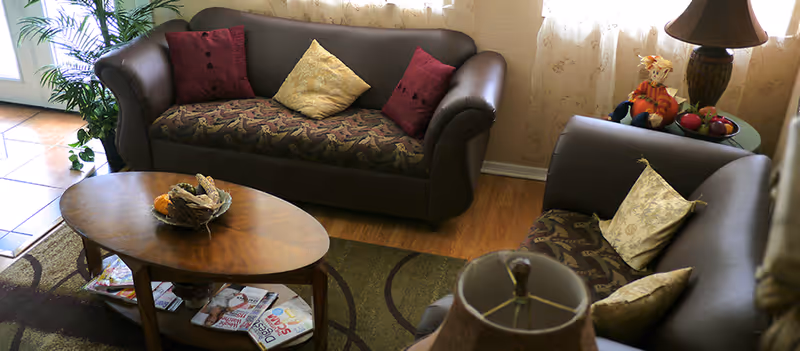 A cozy living room area with two brown sofas adorned with patterned cushions, a wooden oval coffee table with magazines underneath and a decorative centerpiece on top, a side table with a lamp and autumn-themed decorations, and a plant near a tiled floor area.
