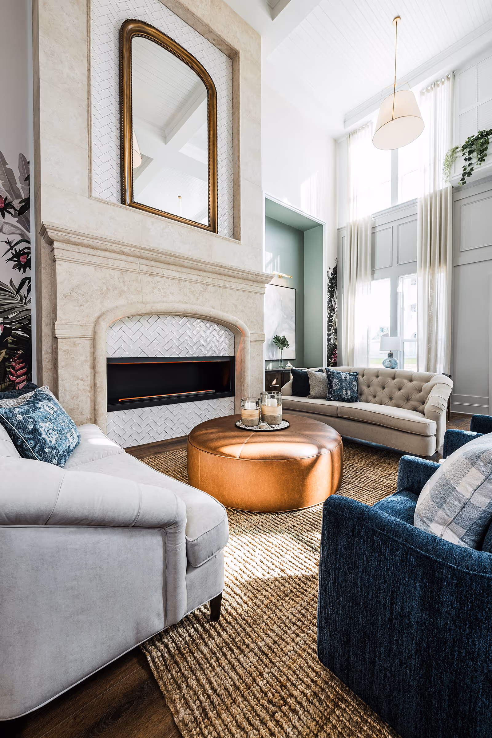 A bright and elegant living room with high ceilings and large windows covered with sheer curtains. The room features a beige tufted sofa, a white armchair with blue patterned pillows, a dark blue armchair with a plaid pillow, and a large round brown leather ottoman in the center. There is a stone fireplace with a large gold-framed mirror above it. The floor is covered with a textured natural fiber rug, and a modern pendant light hangs from the ceiling.