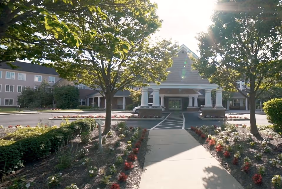 Front exterior view of Horsham Center for Jewish Life showing a paved walkway leading to the entrance with white columns, surrounded by landscaped gardens and trees under bright sunlight.
