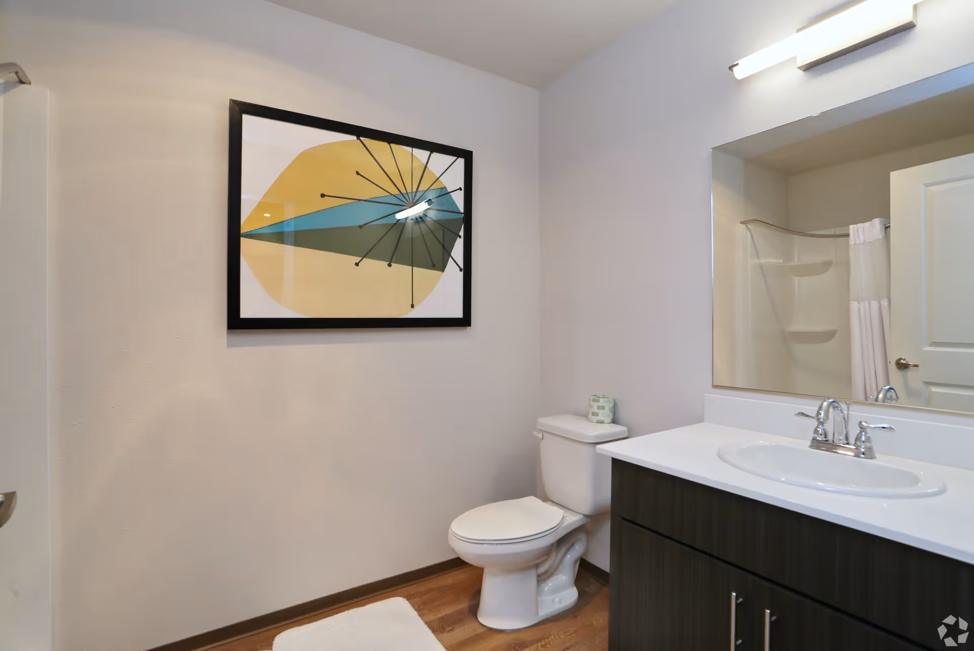 Bathroom with a toilet, vanity and sink, large mirror, shower and framed wall art on a pale wall.