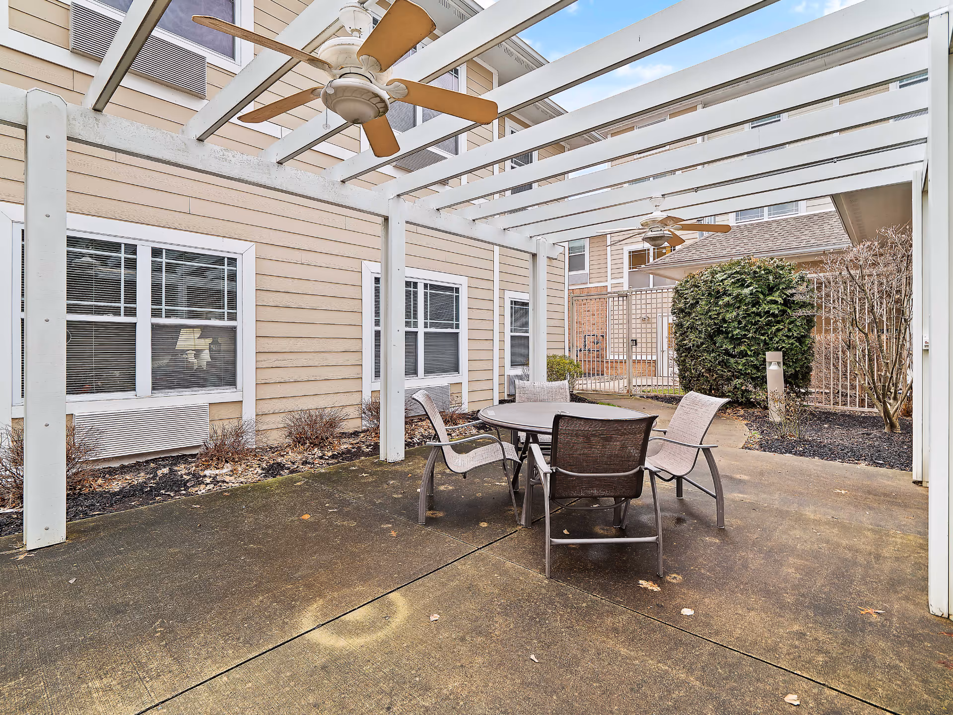 Outdoor patio area with a round table and four chairs under a white pergola with ceiling fans, adjacent to a beige building with windows and some landscaping.