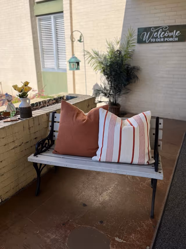 Wood-and-metal bench on a porch with two decorative pillows, potted plants, and a 'Welcome to our porch' sign on the wall.