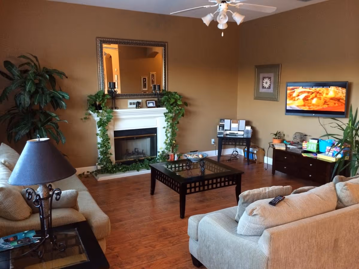 A cozy living room with beige sofas, a glass-top coffee table, a fireplace decorated with green vines, a large mirror above the fireplace, a flat-screen TV mounted on the wall, a side table with a lamp, and various plants and decorative items around the room.