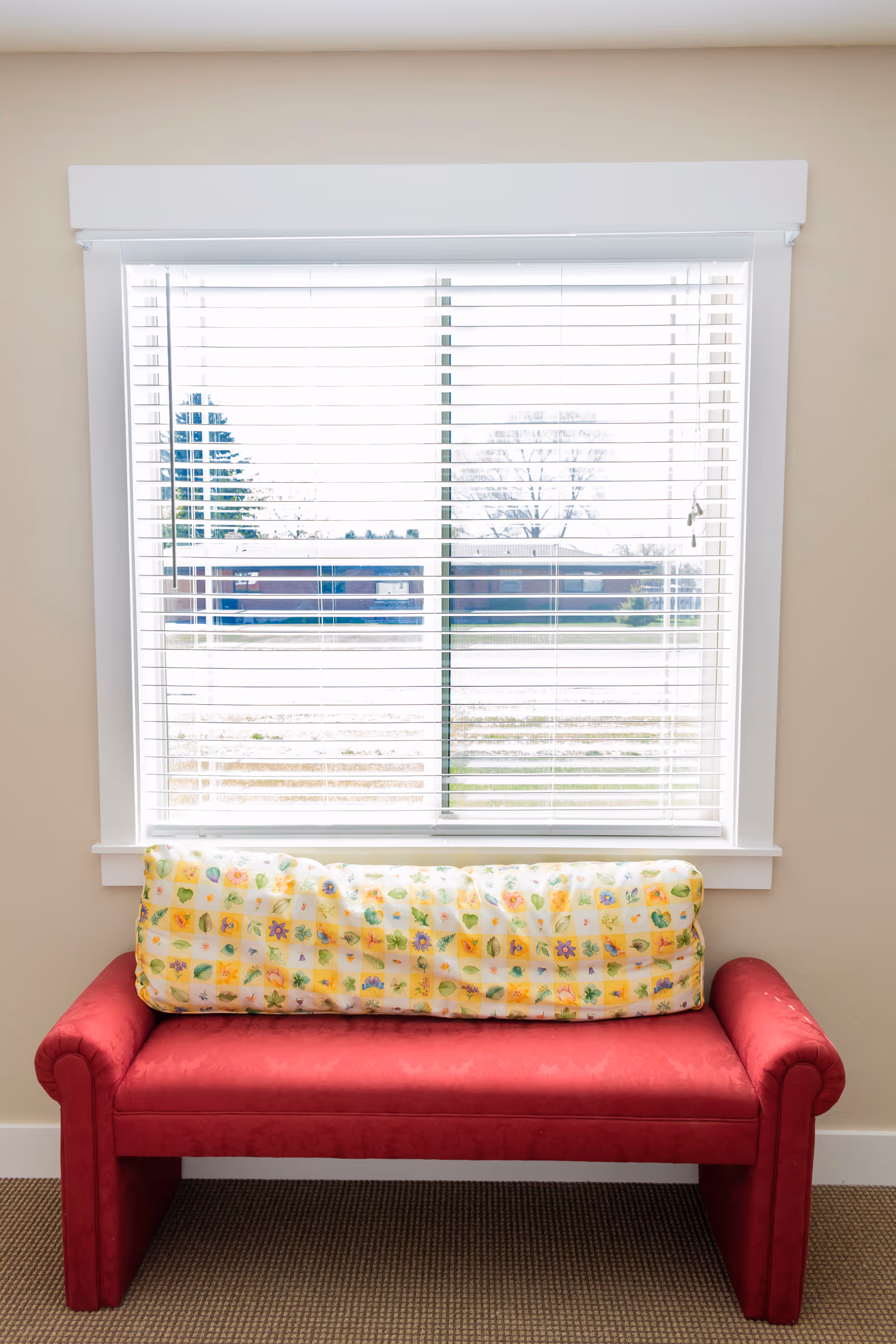 Red upholstered bench with a floral cushion placed beneath a window with white blinds looking out to the street.