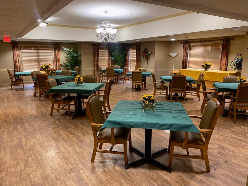 A dining room with multiple tables covered in green tablecloths, wooden chairs, and small sunflower centerpieces on a wood floor.