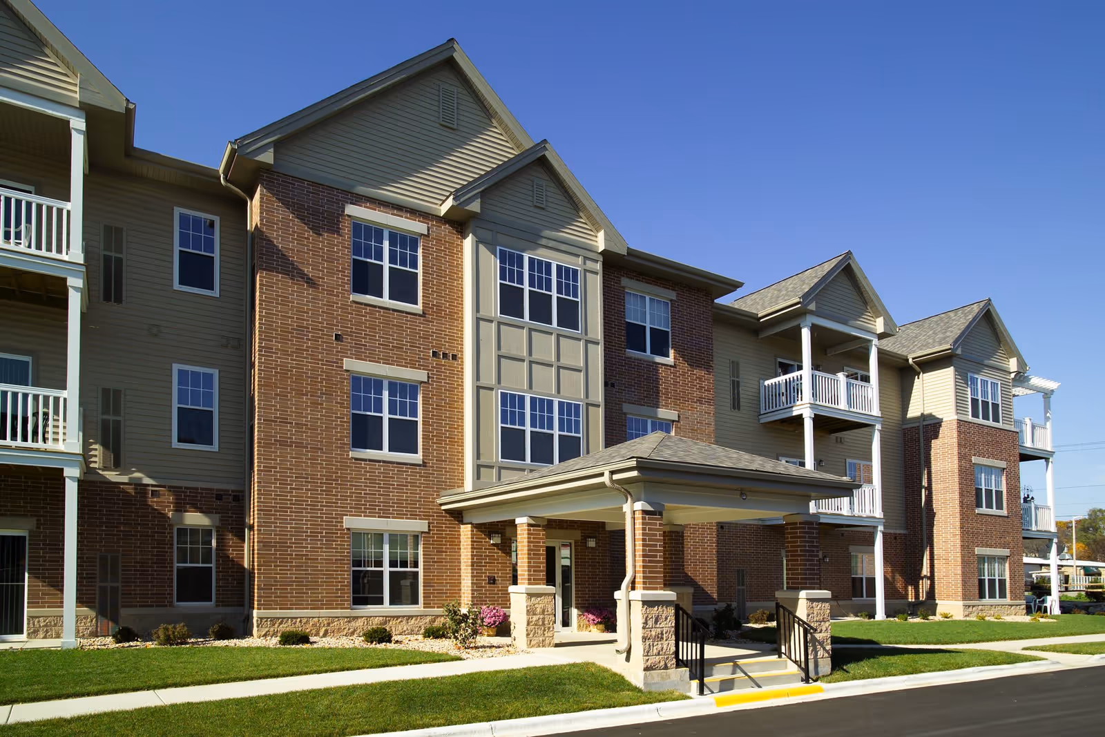 Three-story brick and siding senior living building with a covered entrance, balconies, and a manicured lawn under a clear blue sky.