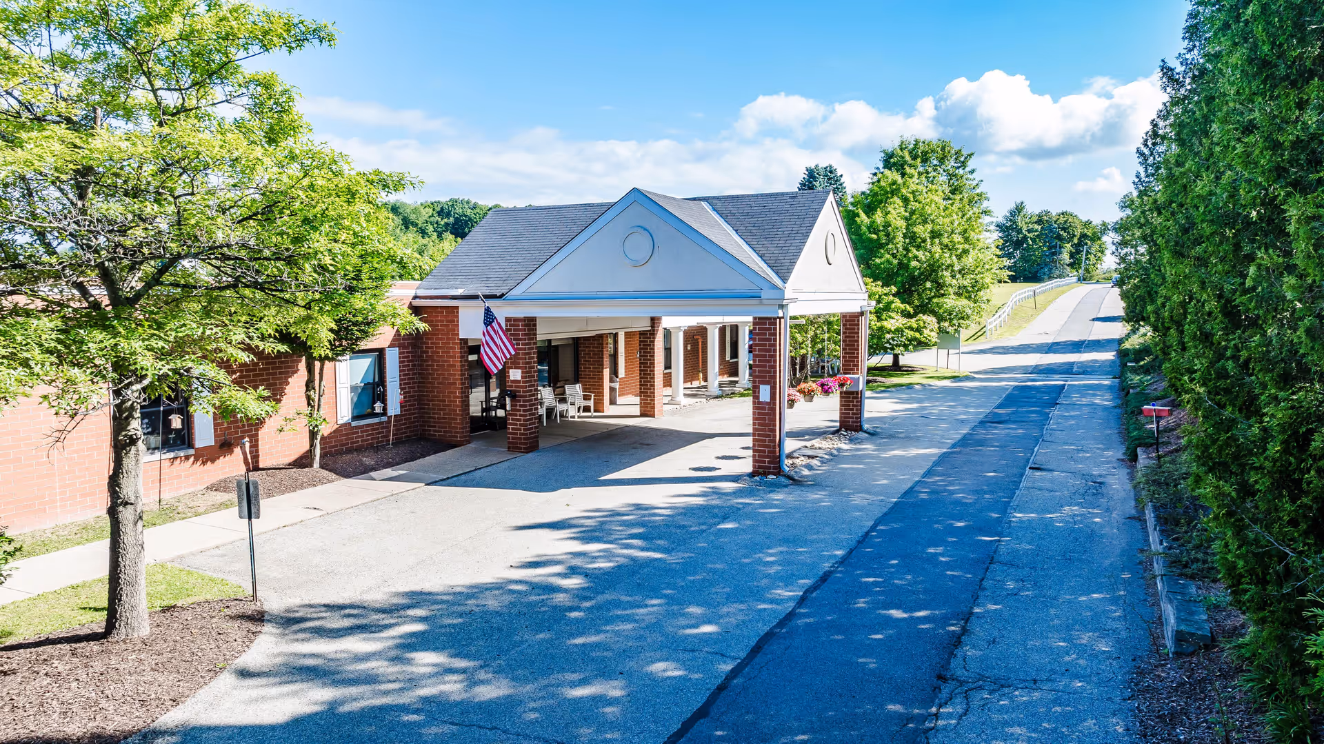 Covered brick entryway with a porte-cochère, American flag, driveway, and trees at the front of a senior care facility.