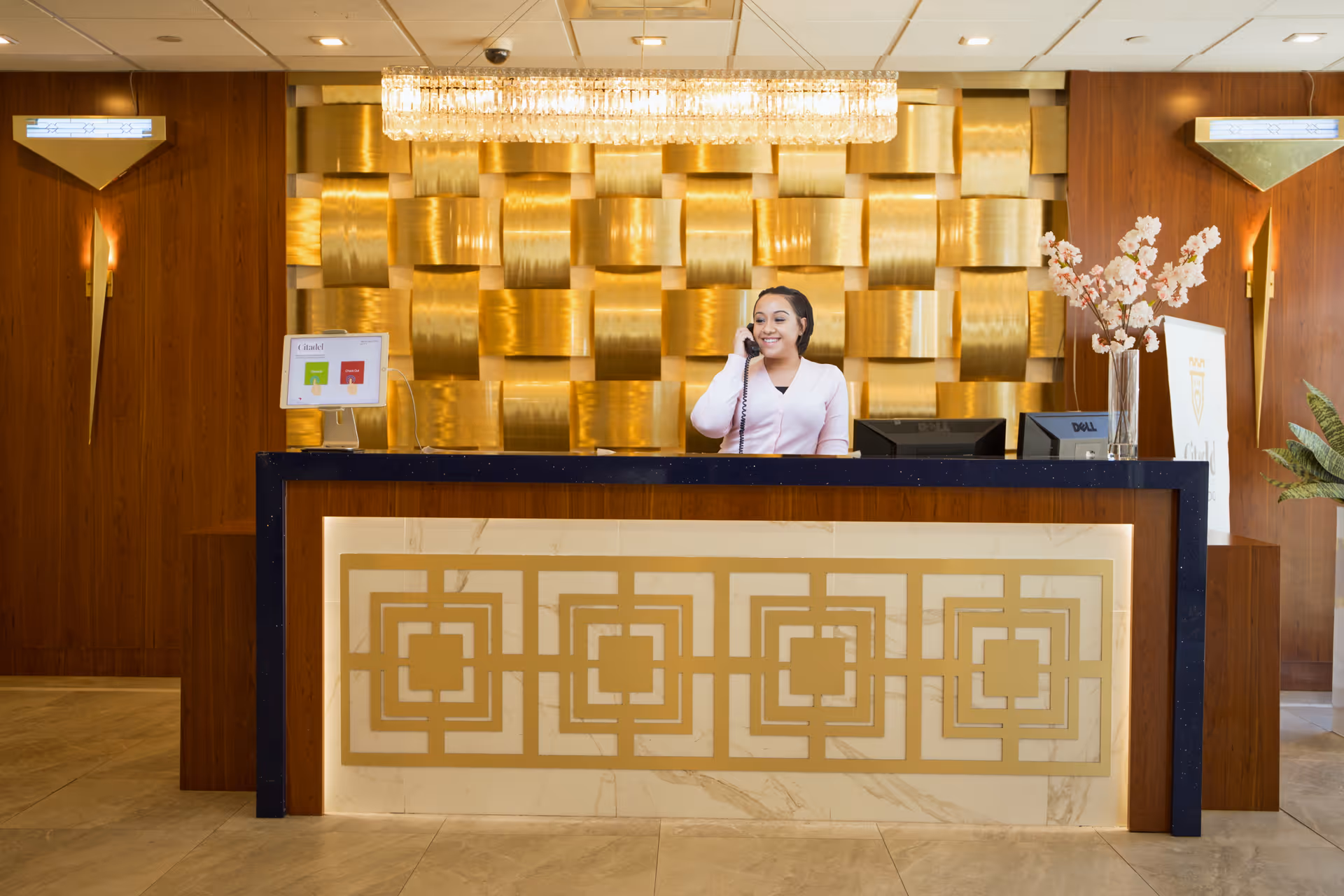 A smiling receptionist talking on the phone behind a decorative front desk with gold geometric patterns. The background features a wall with gold textured panels and a modern chandelier above. There are wooden walls on either side with wall sconces, a vase with pink flowers on the right side of the desk, and a small sign on the left side.