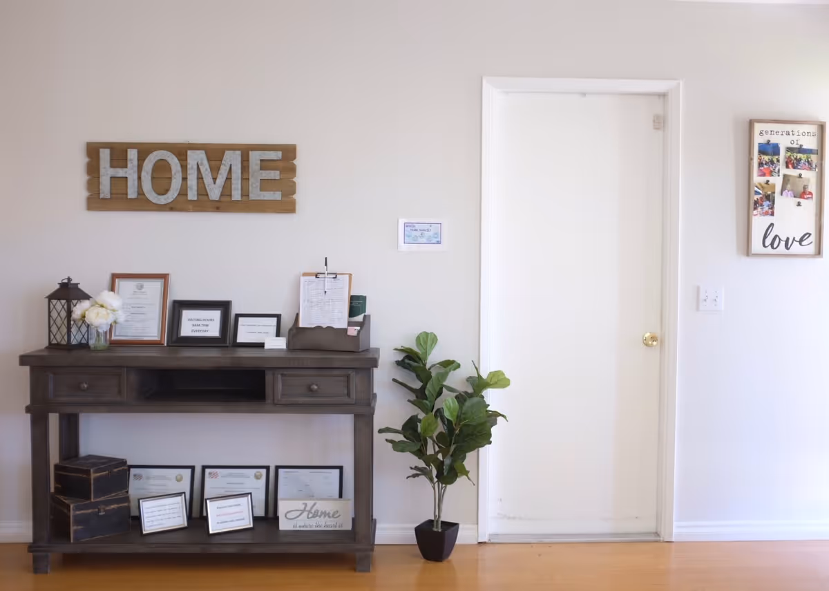 An interior wall of Kamstra Care Home featuring a wooden console table with framed certificates, a lantern, flowers, and informational signs. Above the table is a decorative sign that reads 'HOME'. To the right is a white door and a wall decoration with photos and the words 'generations of love'. A potted plant is placed on the floor next to the door.