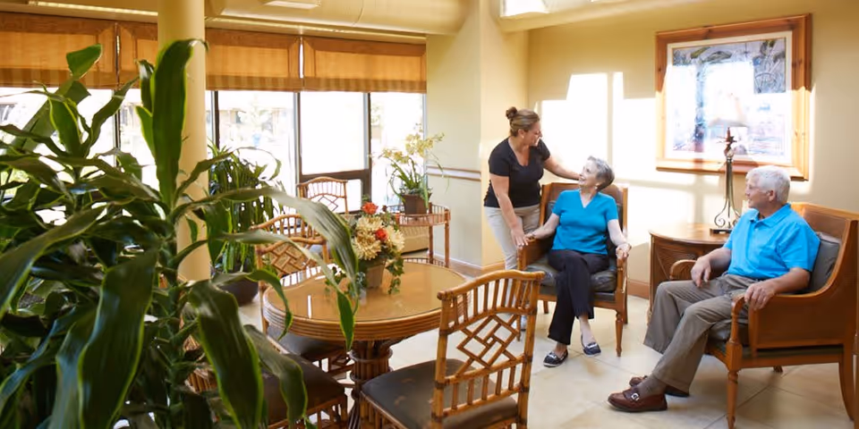 A bright and cozy common area with large windows and natural light. There are wooden chairs and a round table with a floral centerpiece. A caregiver is standing and holding hands with an elderly woman seated in a chair, while an elderly man sits nearby in another chair. The room has plants and framed artwork on the wall.