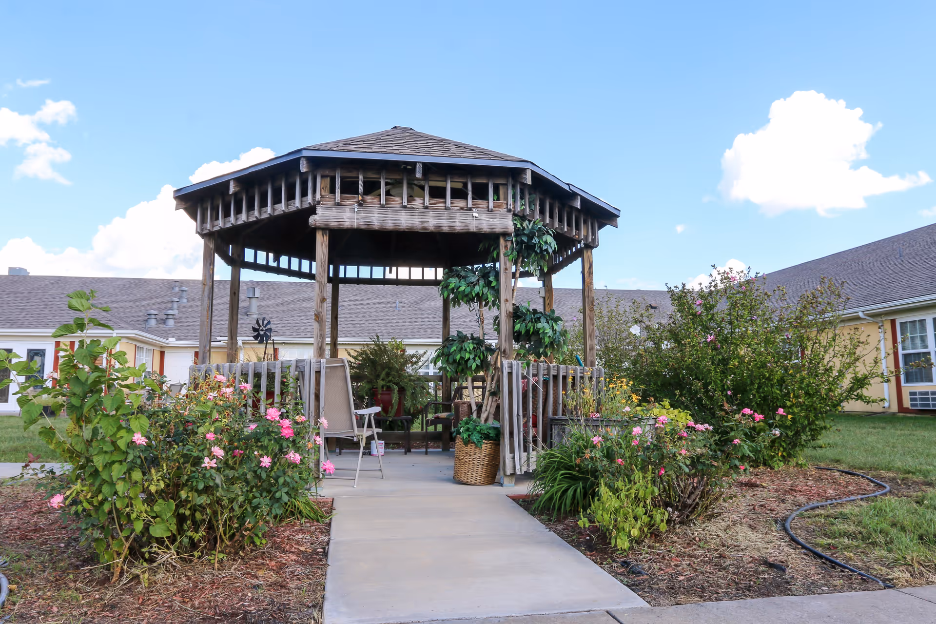 Wooden gazebo with chairs and potted plants surrounded by flower beds and a single-story building under a blue sky.