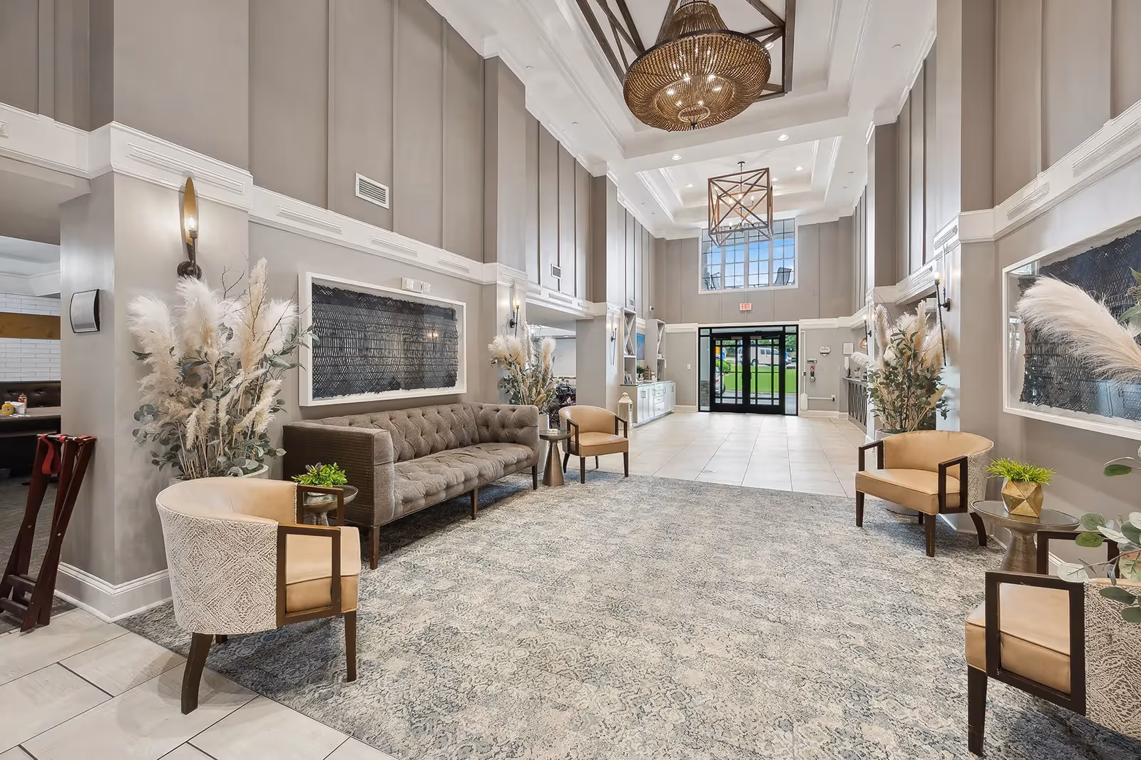 A spacious and elegant lobby area with high ceilings, large windows, and modern light fixtures. The room features a gray tufted sofa, beige armchairs, decorative plants, and a patterned rug. The entrance door is glass with a view of green outdoor space.
