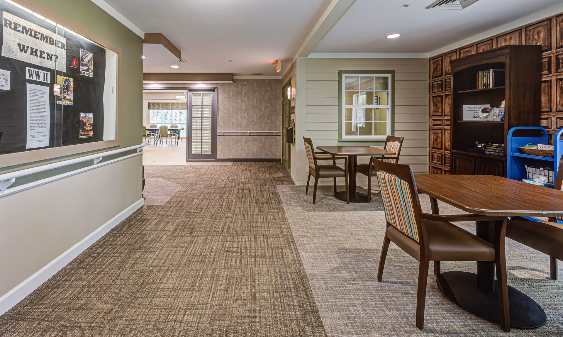 Interior view of a senior living facility hallway with carpeted floors and beige walls. On the right side, there are wooden tables and chairs with striped cushions, a bookshelf, and a blue utility cart. On the left side, there is a bulletin board with historical information and posters titled 'REMEMBER WHEN? WW II'. In the background, a dining area with tables and chairs is visible through an open doorway.