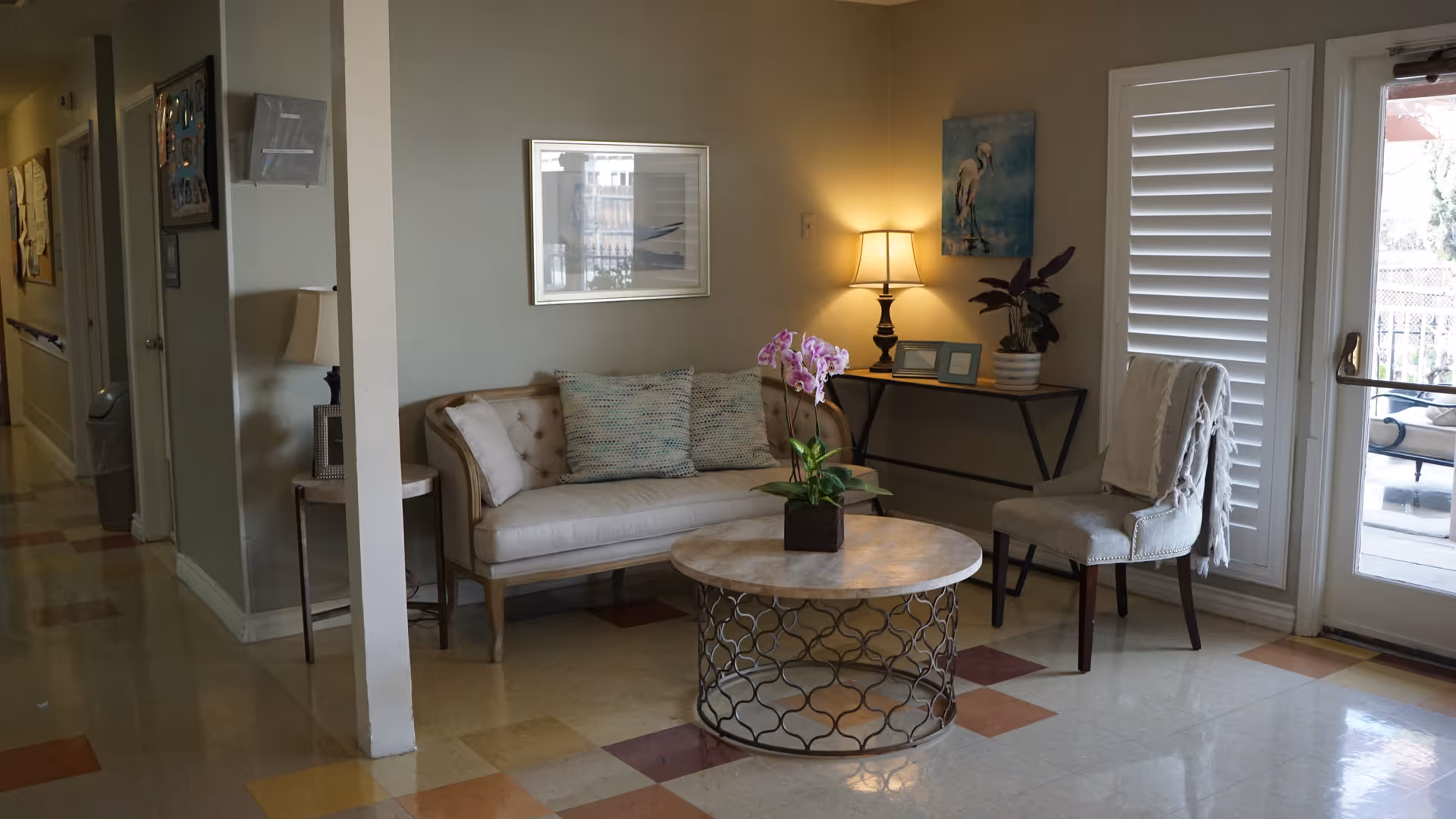 A cozy sitting area in a senior living facility with a beige tufted loveseat adorned with two patterned pillows, a round coffee table with a potted orchid, a cushioned chair with a throw blanket, a console table with a lamp and framed pictures, and a glass door with white shutters leading outside.