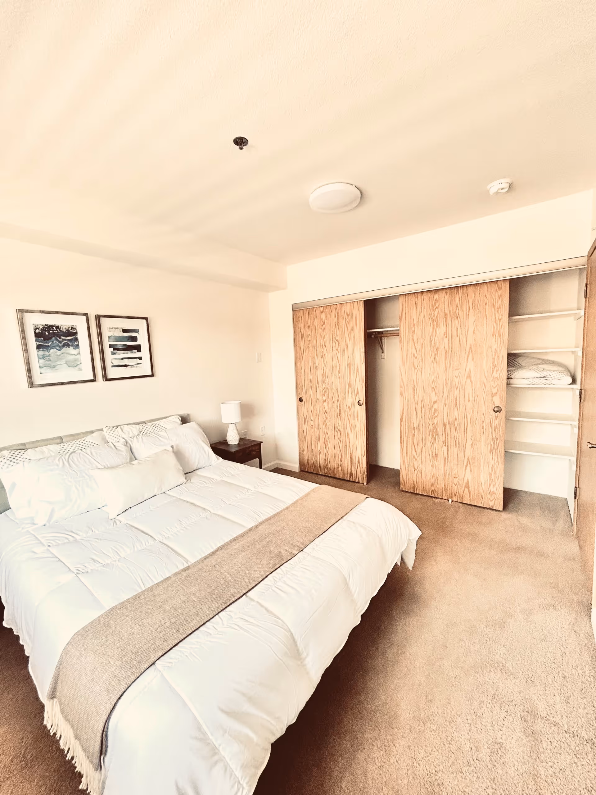 A bright bedroom with a neatly made bed featuring white bedding and a beige throw blanket. There is a wooden nightstand with a white lamp next to the bed. On the wall above the bed are two framed abstract art pieces. Across from the bed is a closet with sliding wooden doors, one door partially open revealing shelves with folded linens.