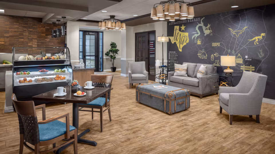 Communal senior living lounge with sofas and armchairs, a trunk-style coffee table, a small dining table with cups, and a refrigerated display of fruit under a mural wall.