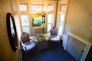 A small sitting area with two wooden chairs with blue cushions facing each other, a square planter with green plants between them, a decorative stained glass window above an air conditioning unit, and a chandelier hanging from the ceiling. The walls are light-colored and there is a large oval mirror on the left wall.