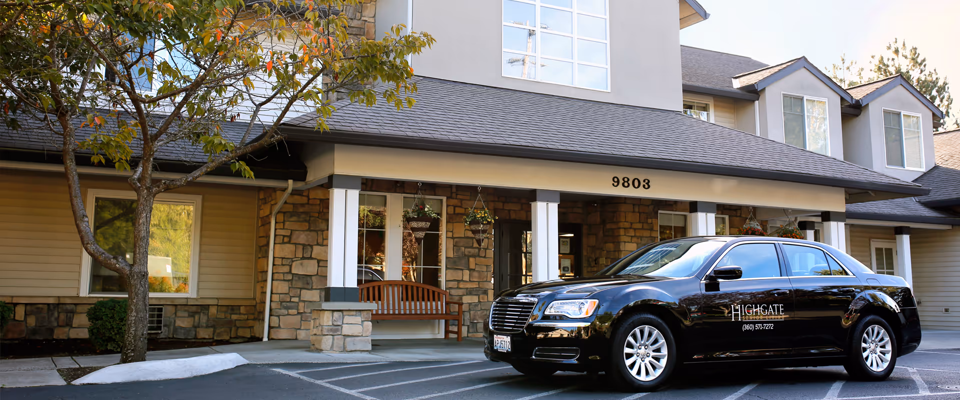Exterior view of Highgate Senior Living - Vancouver building entrance with stone and siding facade, a bench on the porch, and a black sedan car parked in front with the Highgate logo on the door.