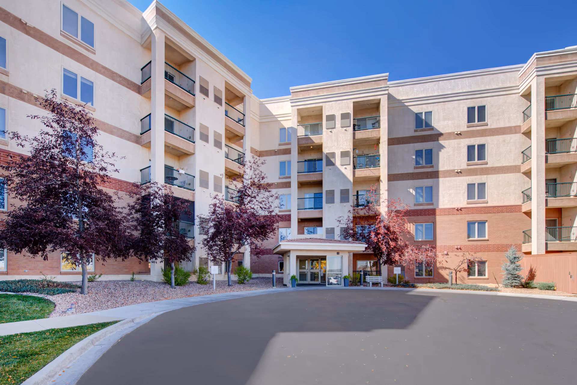 Exterior view of a multi-story senior living facility building with balconies, several trees with purple leaves, and a covered entrance area under a clear blue sky.
