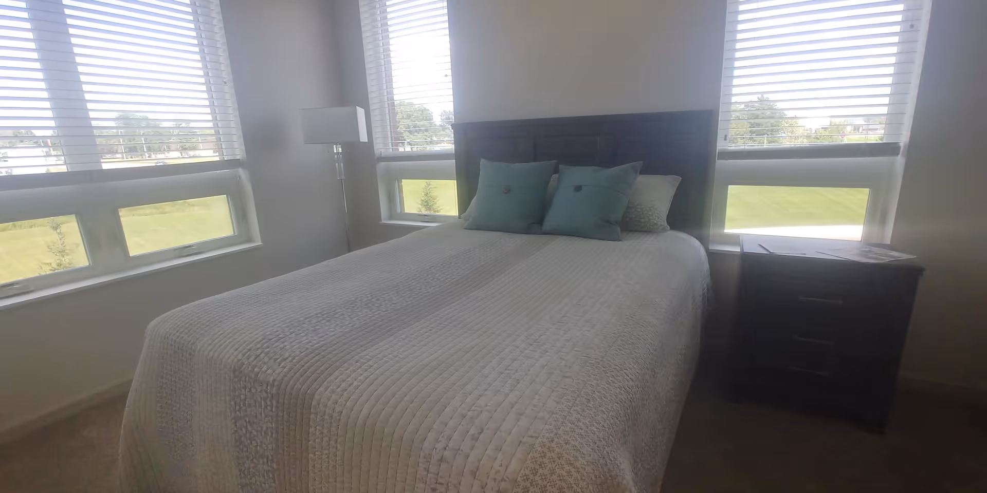 A bedroom with a neatly made bed featuring a white quilted bedspread and three decorative pillows in shades of blue and gray. The bed has a dark wooden headboard. There is a dark wooden nightstand with three drawers to the right of the bed, and a tall floor lamp with a white shade to the left. The room has large windows with white blinds, allowing natural light to brighten the space and offering a view of green grass and trees outside.