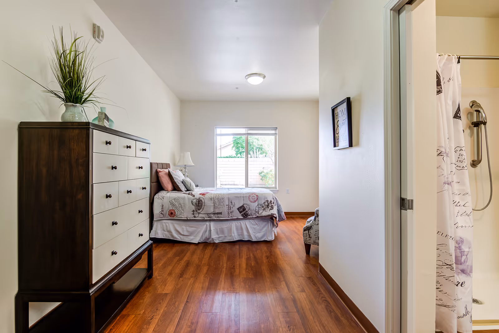 Sunlit bedroom with a bed, tall dresser, hardwood floors and a partially visible bathroom shower.
