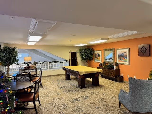 A cozy recreational room with a pool table covered in a yellow cloth at the center. The room has a patterned carpet, orange accent wall with framed artwork, a dartboard, and a wooden cabinet. There are several chairs and tables, a large plant, and a Christmas tree decorated with lights. The ceiling has recessed lighting, and there is a railing overlooking a lower level.