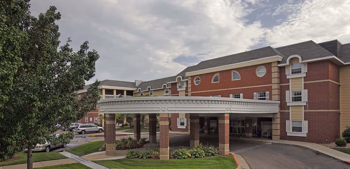 Exterior view of a senior living facility building with a covered entrance supported by brick columns. The building has red brick and beige siding with multiple windows, some with white shutters. There are trees, grass, and parked cars visible around the driveway and entrance area under a cloudy sky.