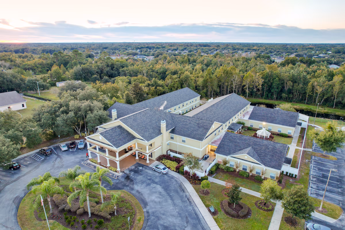 Aerial view of a large L-shaped yellow senior living building with a covered entrance, parking lot, landscaped grounds and surrounding trees.