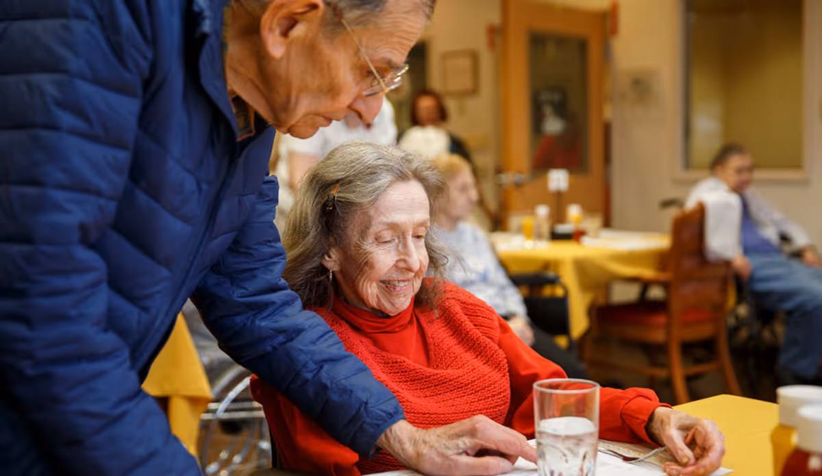 An elderly woman in a red sweater smiles at a dining table as a man leans over to assist her in a communal dining room.