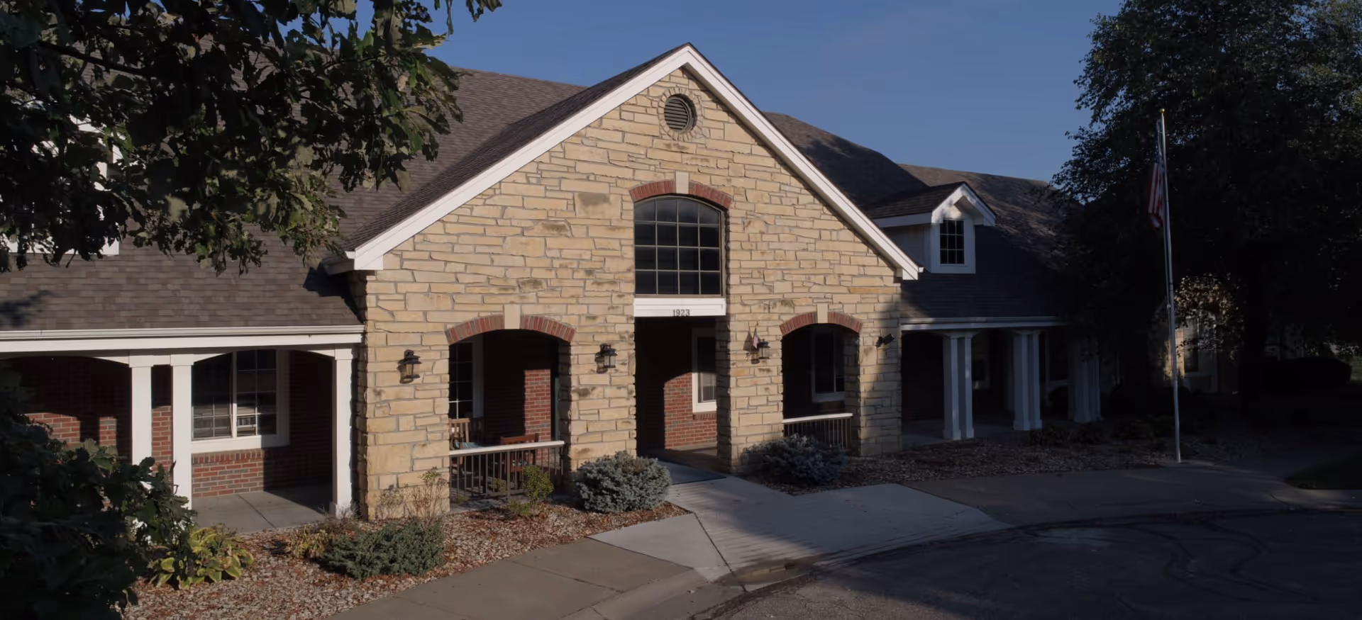 Stone-and-brick assisted living building front with arched entryways, covered porches, landscaping, and an American flag.