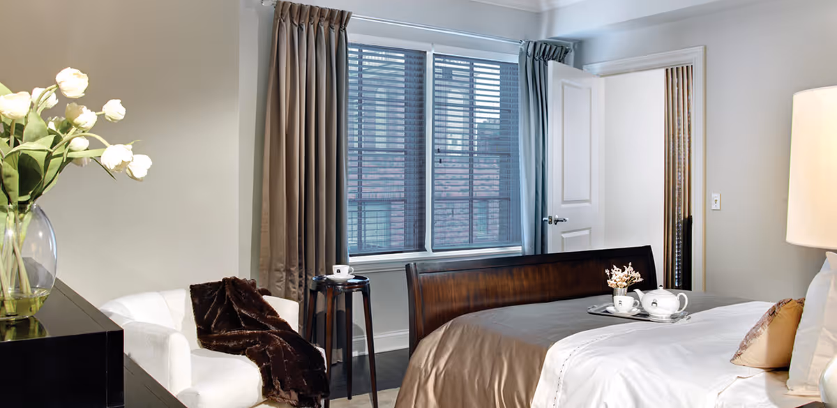 Bright furnished bedroom with a made bed and tea tray, an armchair with a throw, a window with blinds and curtains, and a bedside lamp.